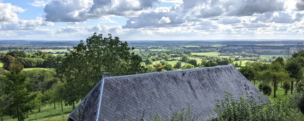 A panoramic view of a serene landscape with rolling hills, dense forest, and a traditional house with a grey slate roof in the foreground, under a partly cloudy sky.