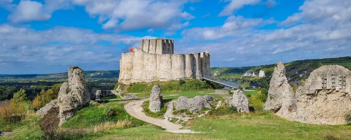 A panoramic view of the ruins of Chateau Gaillard, an ancient fortress situated on a hilltop surrounded by lush green landscapes under a partly cloudy sky.