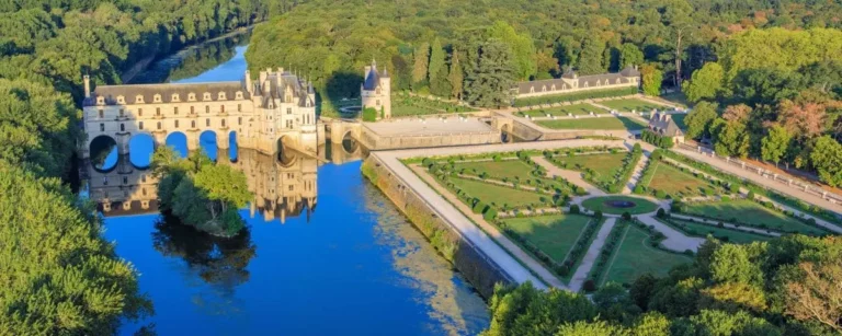 Aerial view of Chenonceau Castle with its distinctive arches over a river, surrounded by manicured gardens and dense forest.