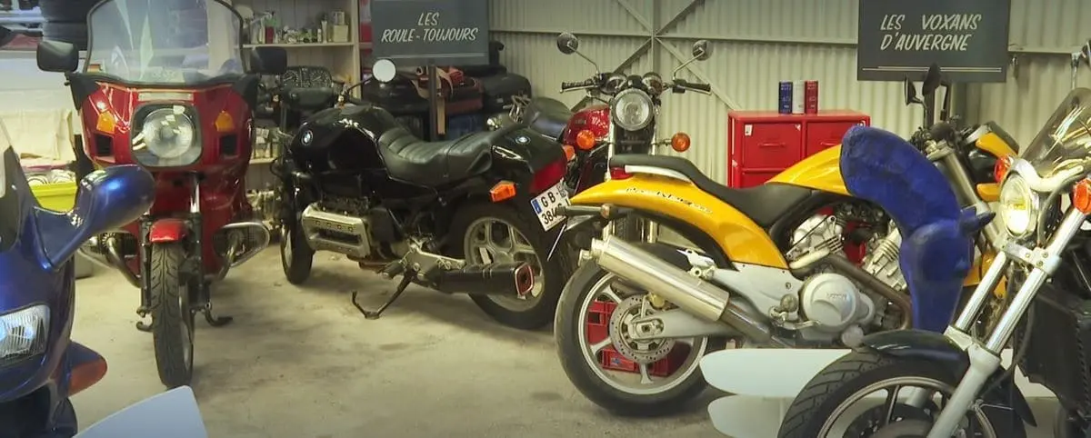 A collection of vintage motorcycles displayed in a garage setting at the Chimomu Museum in Chinon. Signs in French indicate different motorcycle groups.