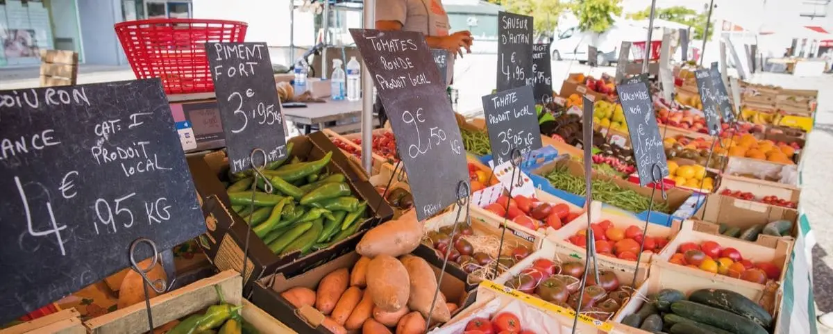 A vibrant outdoor food market in Chinon showcasing various fruits and vegetables. Handwritten price signs in French are visible among the produce.