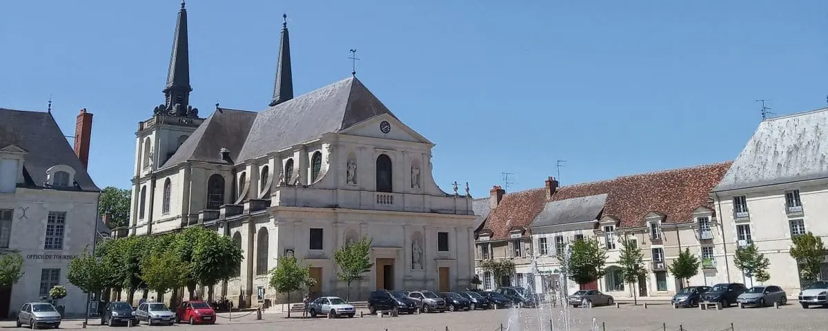 The historic Church of Notre-Dame in Richelieu, featuring two tall spires, surrounded by quaint buildings and a cobblestone square with a fountain.