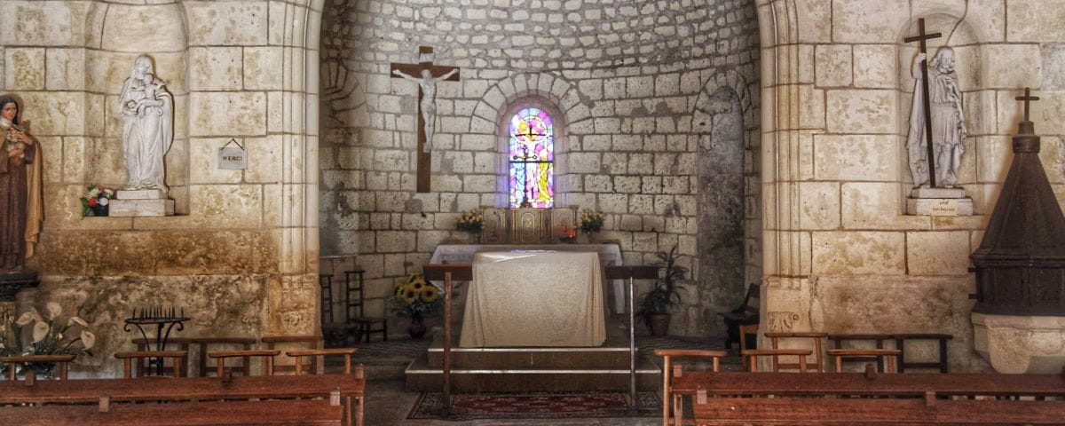 The interior of the Church of Saint-Jean-Baptiste in Chenonceaux, featuring a stone altar adorned with a white cloth, a crucifix, and statues on either side. The walls are made of stone, and there's a stained glass window above the altar.
