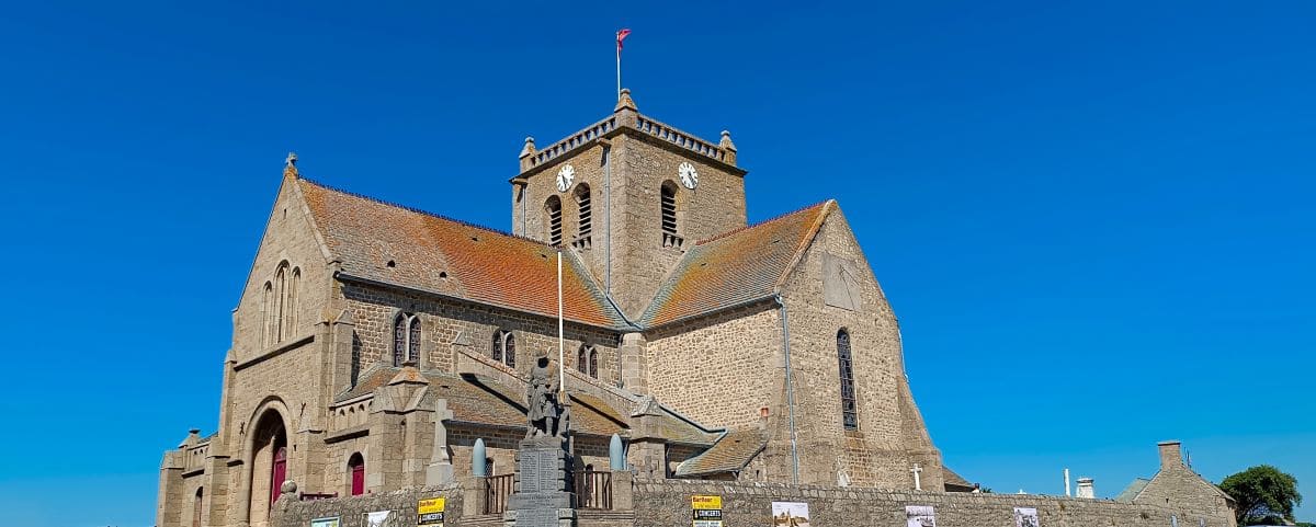 The Church of Saint Nicolas in Barfleur, a historical stone church with a clock tower and red-tiled roof, set against a bright blue sky. The church features arched doorways and windows.