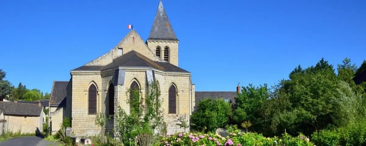 A quaint stone church in Montsoreau with a pointed steeple and surrounding greenery, set against a clear blue sky.