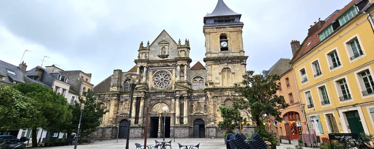 The façade of the Church of Saint-Rémy in Dieppe, featuring detailed stonework, a clock tower, and surrounding colorful buildings under a cloudy sky.