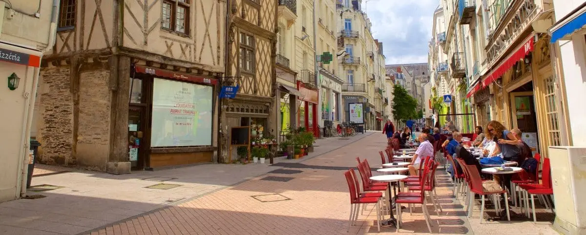 Busy city street lined with historic buildings, outdoor café seating, and people walking and dining under a clear sky.