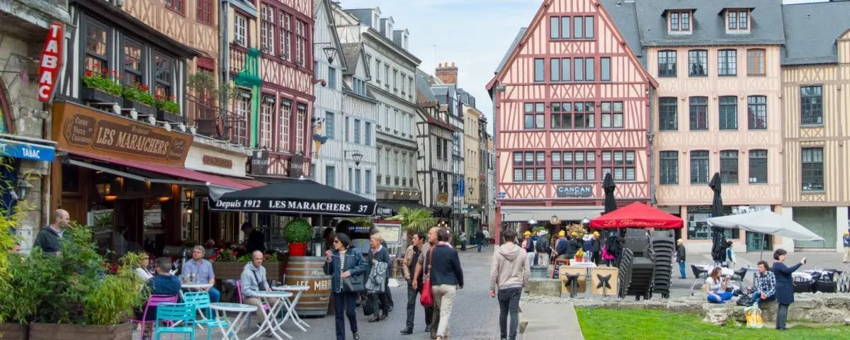 A lively scene in the city center of Rouen, featuring people dining at outdoor cafes and strolling along the street, lined with colorful, half-timbered buildings housing shops and restaurants.