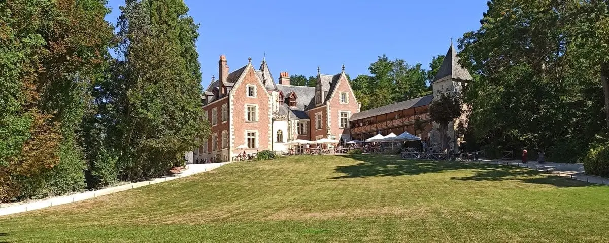 A picturesque view of the Clos Lucé, a historic mansion with red brick and stone architecture surrounded by lush greenery and a well-manicured lawn. The mansion is a significant site related to Leonardo da Vinci
