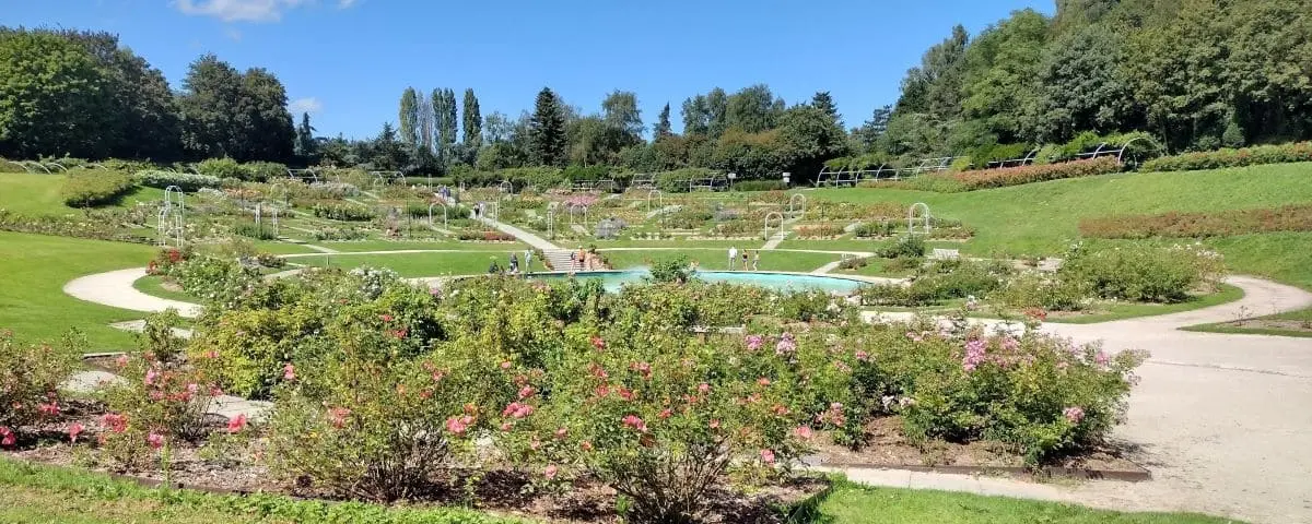 A beautifully landscaped park with blooming flower beds and a central fountain. Pathways wind through the greenery, inviting visitors to explore the serene environment.