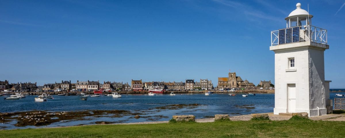 The Cracko Lighthouse in Barfleur, a white lighthouse overlooking a calm harbor with moored boats. The distant shore shows the quaint town of Barfleur with stone buildings and a church, all under a clear blue sky.