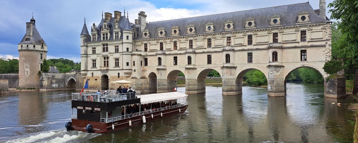 A boat cruise on the Cher River passing in front of the Château de Chenonceau, with the castle's iconic arches over the water. The boat is filled with tourists, and the sky is partly cloudy.