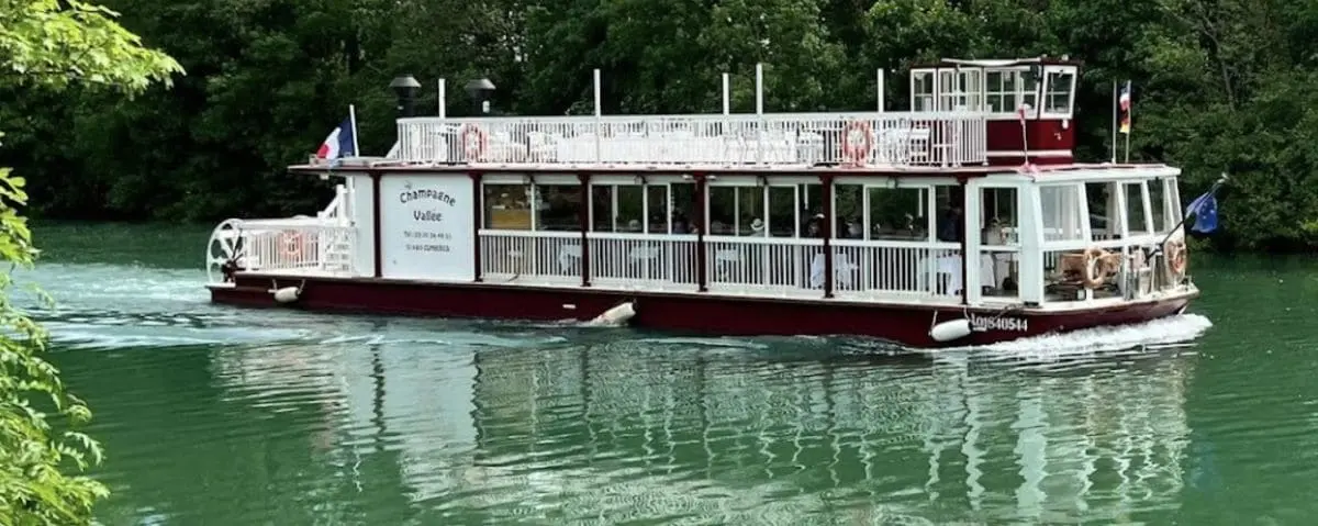 A red and white boat named 'Champagne Vallée' cruising along the serene Marne River surrounded by green foliage.