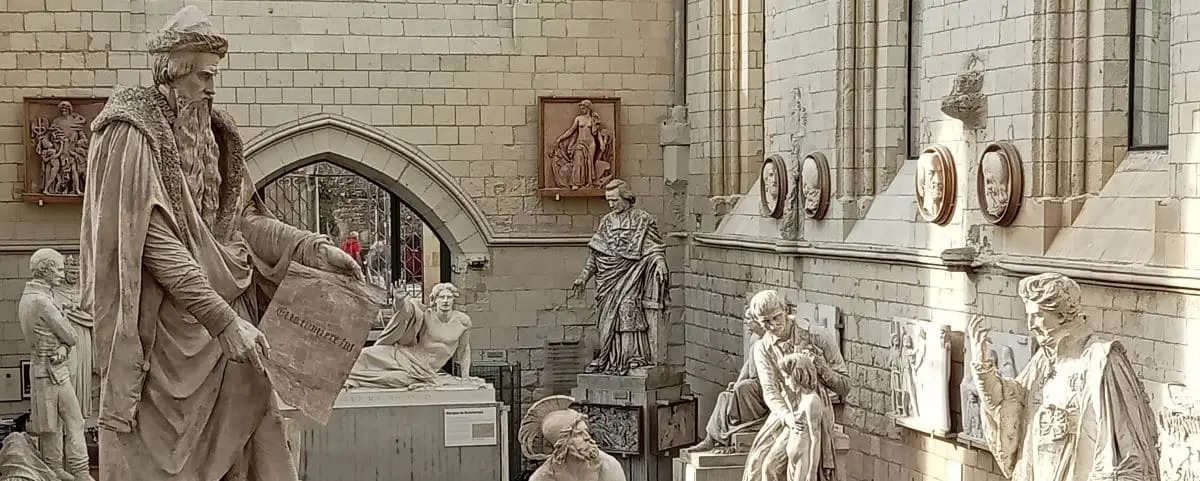 Gallery room filled with stone statues and sculptures, situated in a Gothic-style building with arched windows and stone walls.