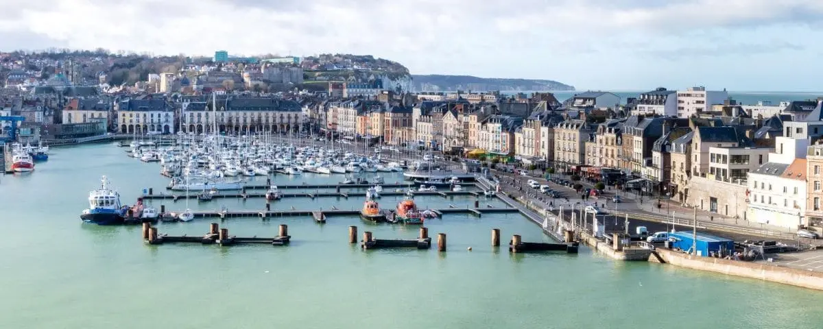 Scenic view of Dieppe town with a marina filled with boats, historic buildings along the waterfront, and a backdrop of hills and a cloudy sky.