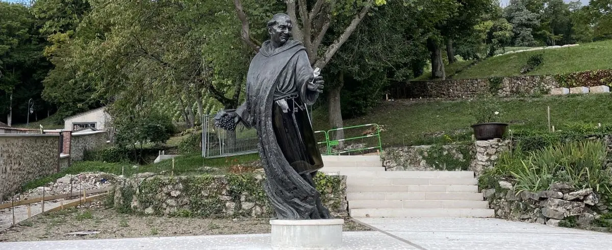 A statue of Dom Pérignon in Hautvillers, depicting him holding a bunch of grapes, surrounded by greenery and stone steps leading up to the monument.