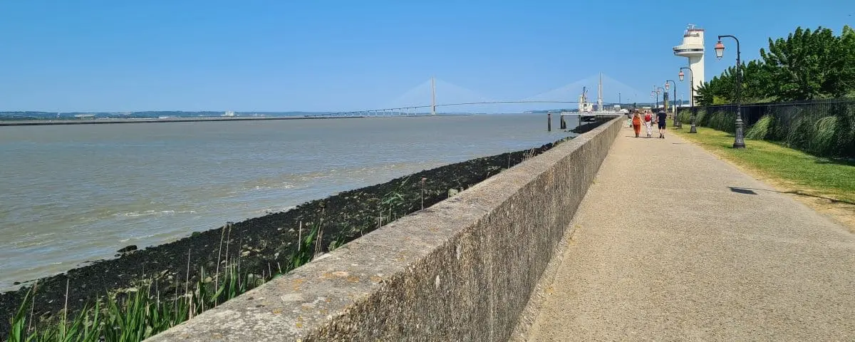 A view along the dyke in Honfleur, with a concrete walkway next to the water. In the background, the Pont de Normandie bridge can be seen spanning the water.