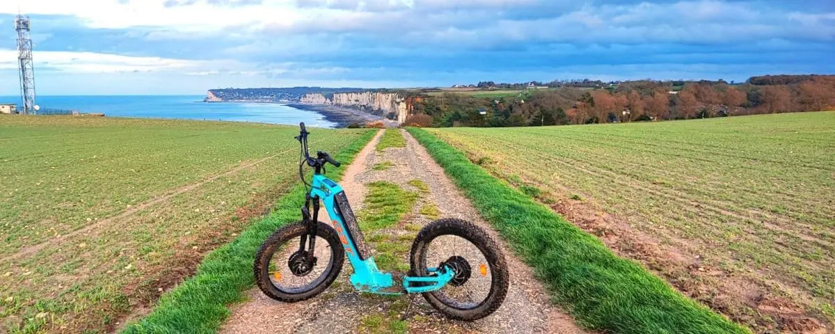 A blue electric scooter on a path overlooking the coastal cliffs and countryside of Fécamp, with the sea visible in the distance.
