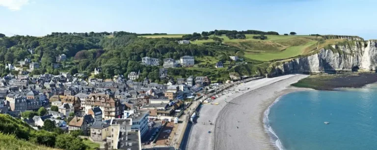 Panoramic view of Etretat town with its pebble beach, white cliffs, and lush green hills, all overlooking the clear blue sea.