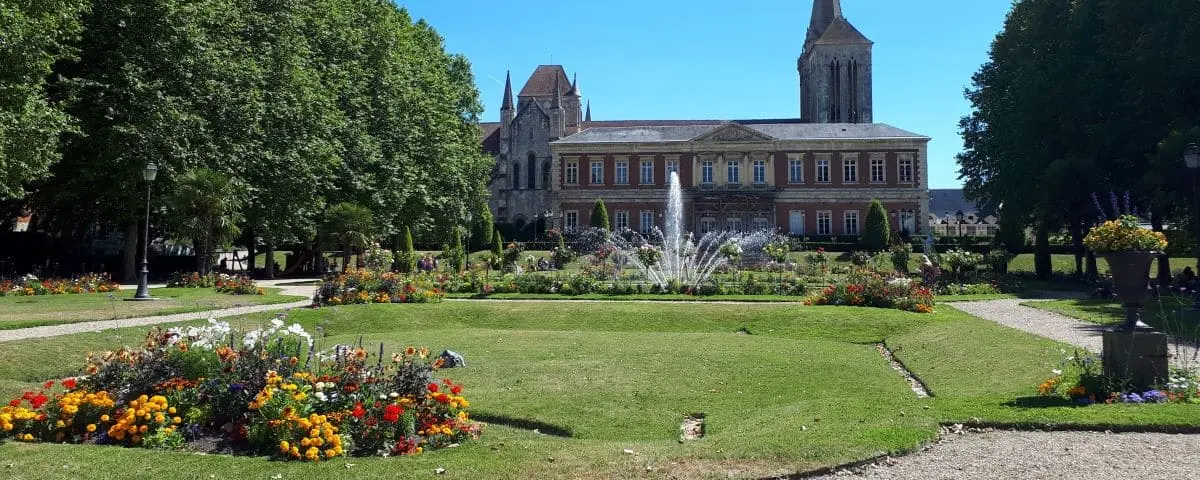 A beautifully landscaped garden with vibrant flower beds and a central fountain, situated in front of a grand historic building. The garden is surrounded by lush green trees and neatly maintained paths.