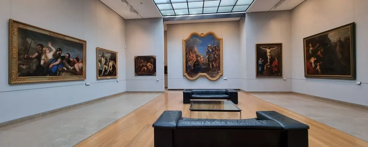 An interior view of a gallery in the Fine Arts Museum, displaying large framed paintings on the walls. Comfortable seating areas are placed in the center for visitors to admire the artwork.