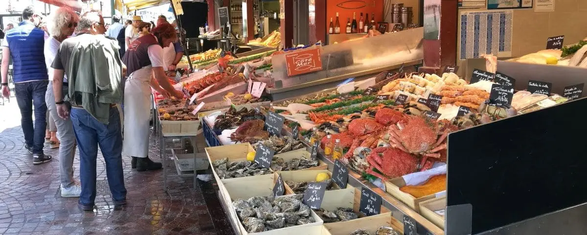 A bustling fish market in Trouville with various seafood displayed, including crabs, oysters, and lobsters. People are seen selecting and buying fresh seafood.