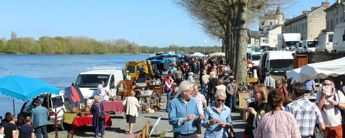 A bustling flea market along the riverside in Montsoreau, with vendors selling various items under tents and people browsing in the bright sunshine.