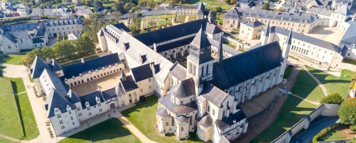 Aerial view of Fontevraud Abbey showcasing its large, historic complex of stone buildings with dark roofs, surrounded by green lawns and nearby town structures.