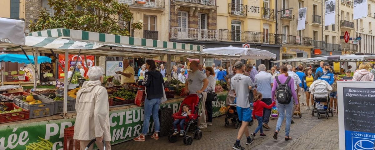 A bustling market scene in Dieppe with people shopping at various stalls selling fresh produce under striped awnings. The market is set against a backdrop of historic buildings.