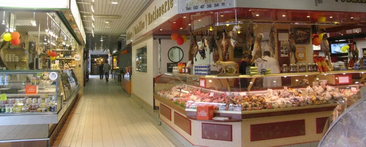 An indoor food market in Tours, France, with various stalls selling meats, cheeses, and other produce, with bright lighting and a few shoppers walking through.