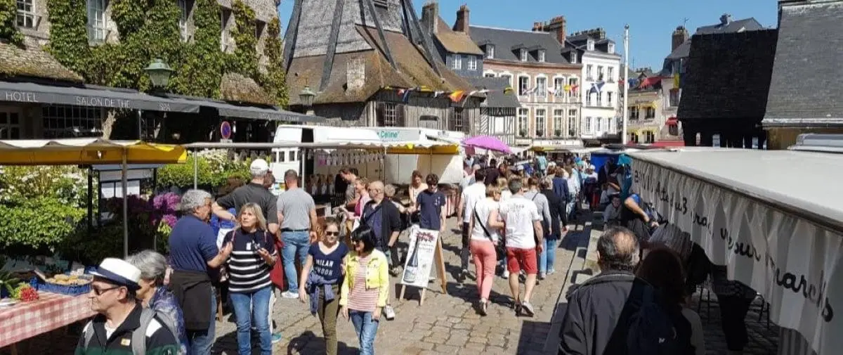 A bustling outdoor food market in Honfleur, with people walking among various stalls. The market is lively with a variety of vendors and shoppers enjoying the vibrant atmosphere.