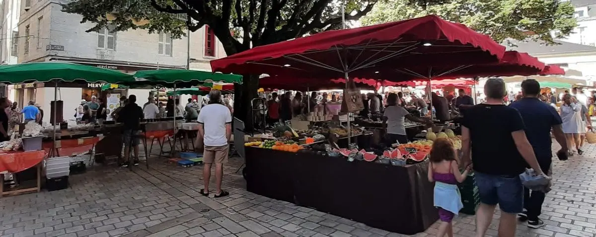 A bustling outdoor food market in Périgueux, with stalls covered by red and green canopies. Shoppers browse a variety of fresh produce and local goods, contributing to a lively and vibrant atmosphere.