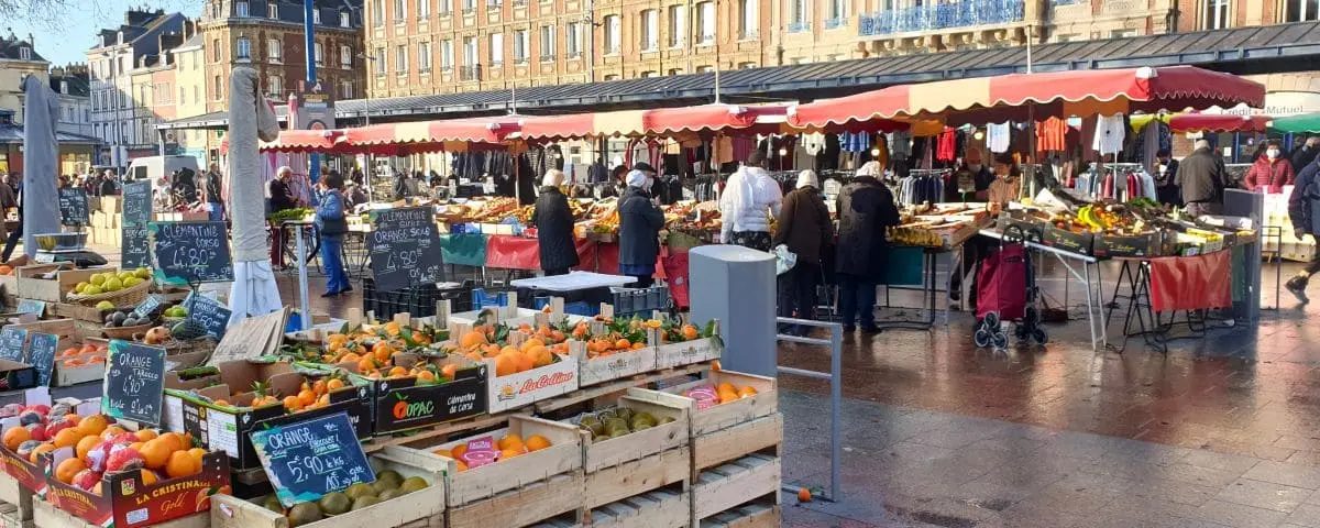 A bustling food market in Rouen, with stalls selling a variety of fresh produce, including oranges and other fruits, and people browsing and shopping under red and white awnings.