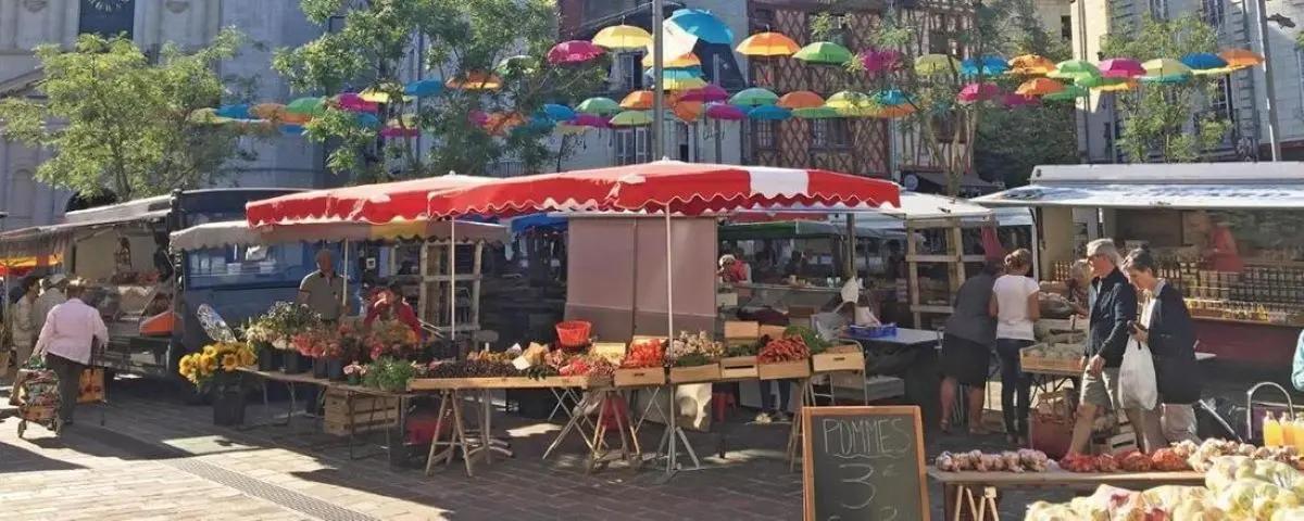 Vibrant outdoor food market in Saumur with colorful umbrellas overhead, stalls selling fresh produce and flowers, and shoppers browsing the goods.