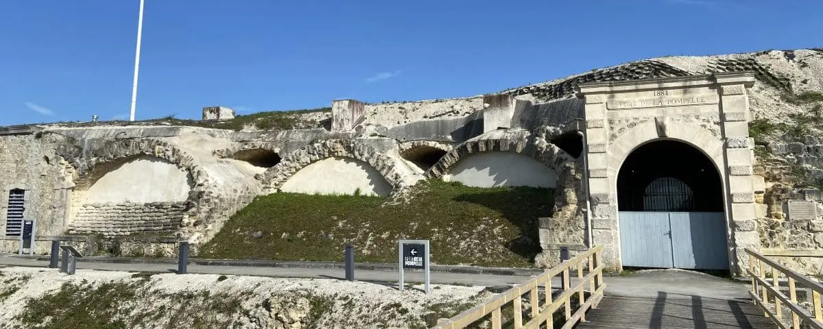 The entrance to Fort de la Pompelle Museum, featuring arched stone structures and a historical plaque, under a clear blue sky.