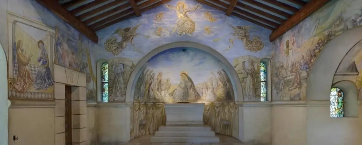 Interior view of the Foujita Chapel in Reims, featuring intricate wall paintings and a vaulted ceiling decorated with religious artwork. The serene chapel is adorned with stained glass windows and a central altar.