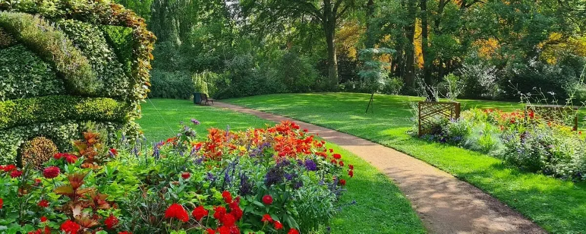 A vibrant garden at Pierre Schneiter Park in Reims, showcasing a colorful array of flowers and lush greenery along a winding path. The well-maintained garden is surrounded by tall trees and serene natural beauty.