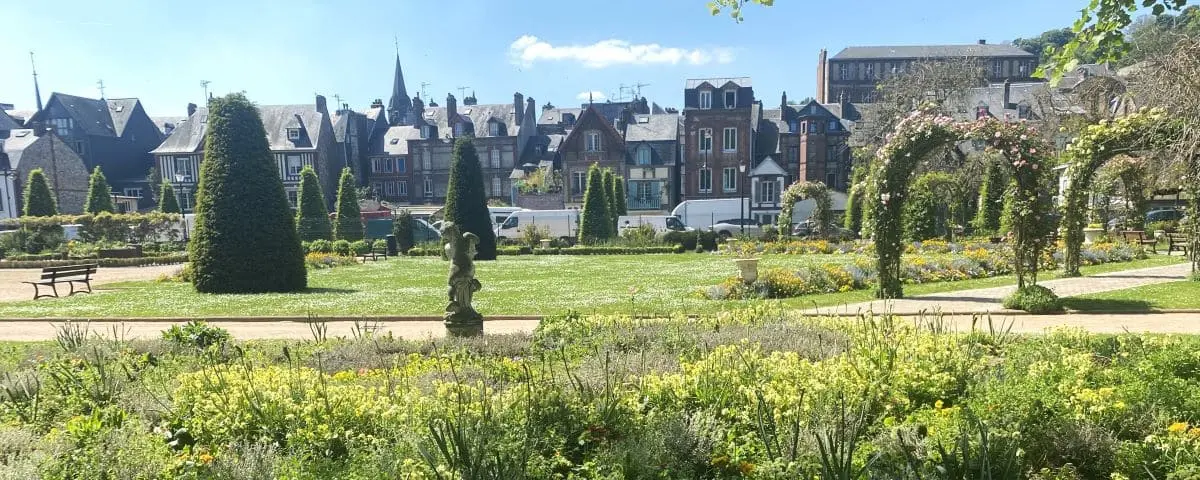 A beautifully landscaped garden with neatly trimmed hedges, colorful flower beds, and a variety of plants. The background shows charming buildings with a mix of architectural styles.