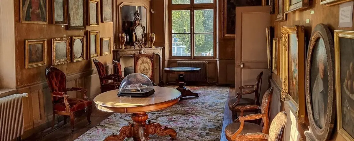 An elegantly decorated room in a museum, filled with vintage furniture, framed paintings, and a display case in the center. The atmosphere is warm and inviting.