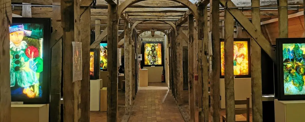 An exhibition at the Gemmail Museum in Montrésor displaying vibrant, illuminated glass artworks in a rustic, wooden gallery setting.