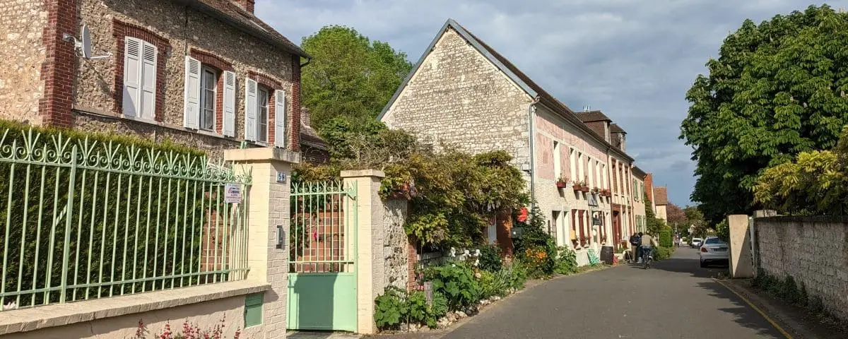 A quaint street in Giverny with charming stone houses, green shutters, and lush greenery. The quiet scene includes a couple of people walking or cycling under a partly cloudy sky.