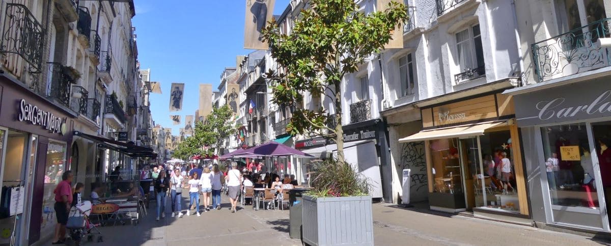 The busy Grande Rue in Dieppe, lined with shops and cafes, with people walking and dining outdoors on a sunny day. Banners are strung across the street, adding to the lively atmosphere.