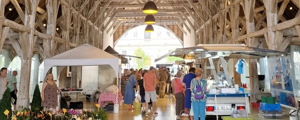 A bustling food market in Richelieu under a large wooden hall, with vendors selling fresh produce and local goods, and people shopping.