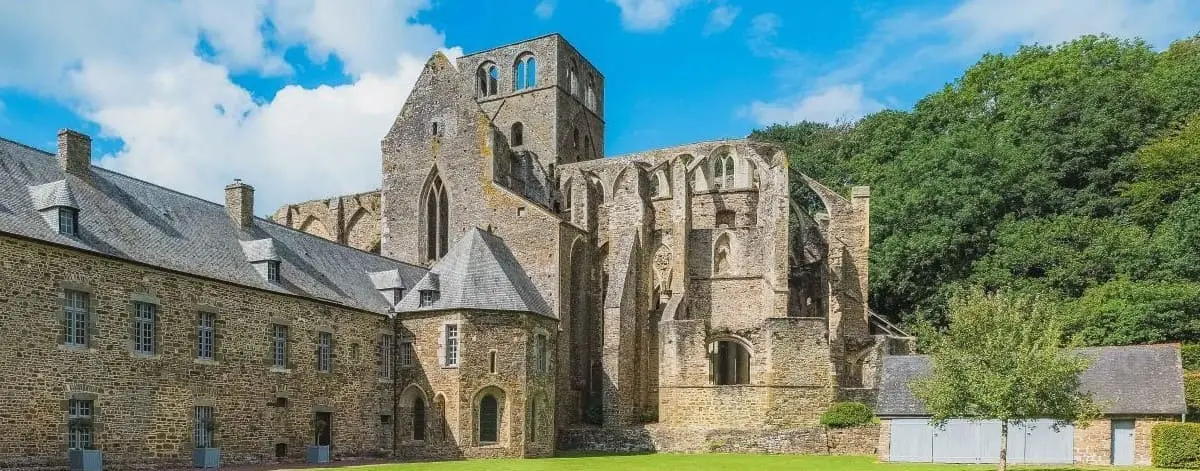 Ruins of Hambye Abbey, showcasing its medieval stone architecture with partially intact walls and arches set against a backdrop of green lawns and trees.