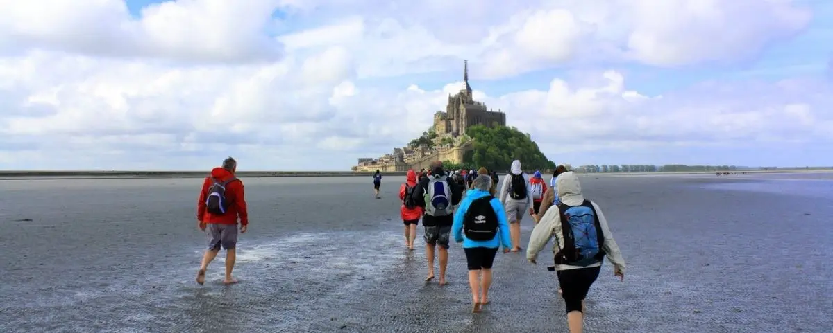 A group of people hike barefoot across the sandy bay towards Mont Saint-Michel under a partly cloudy sky.