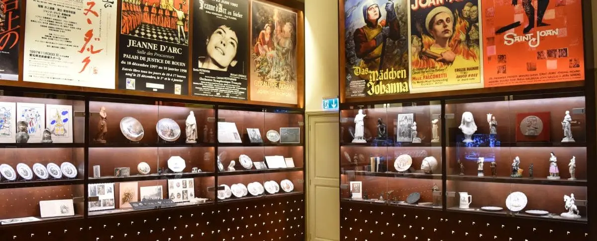 A display at the Historial Jeanne d’Arc in Rouen, featuring posters of Joan of Arc films and various memorabilia, including plates and statues dedicated to her legacy.