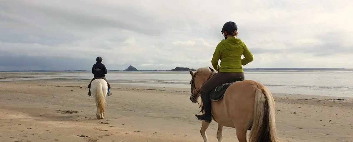 Two riders on horseback travel along the beach with Mont Saint-Michel visible in the distance against a cloudy sky.