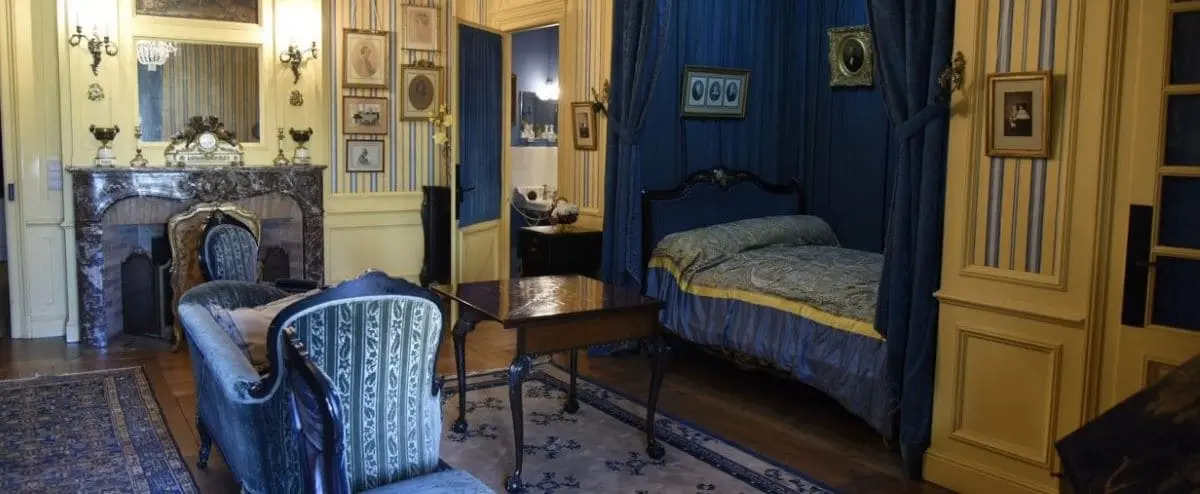 A historical room at the Hotel Museum Le Vergeur in Reims, featuring elegant antique furniture, a blue canopy bed, and ornate decor. The room exudes old-world charm with framed portraits and a grand fireplace.