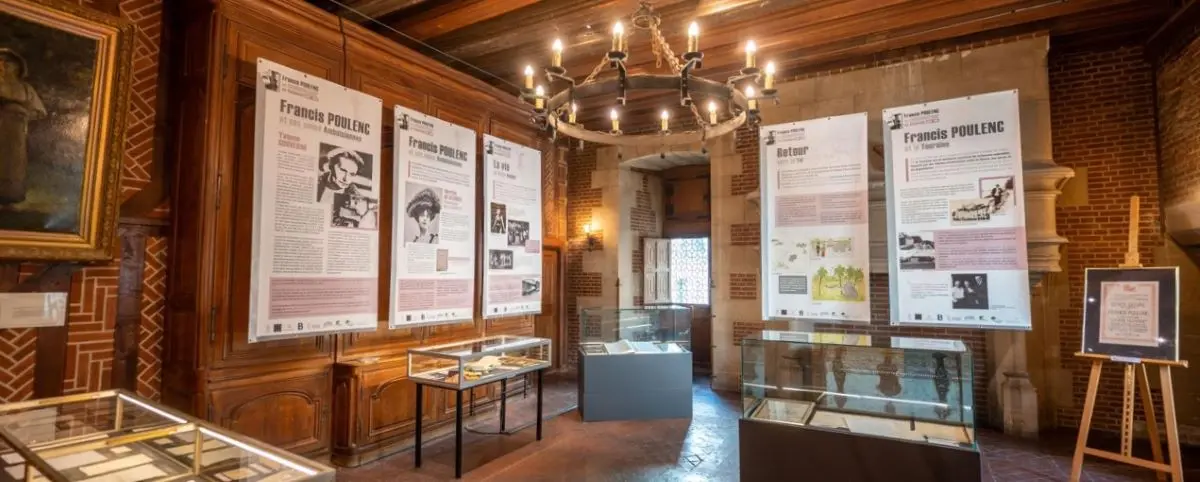 An interior view of the Hotel Museum Morin, showcasing exhibits dedicated to Francis Poulenc. The room features informative displays, glass cases with artifacts, and a rustic wooden chandelier hanging from the ceiling.