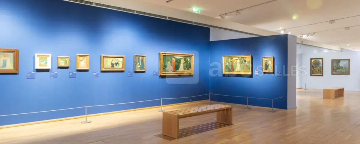An exhibition room in the Impressionism Museum with blue walls displaying framed impressionist paintings. The room features wooden flooring and benches for visitors to sit and observe the artwork.